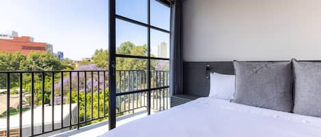 A modern hotel room with a large bed, a balcony with a view of the city, and a large window.
