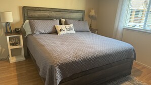 Ein ordentlich bezogenes Bett mit einer grauen Decke, zwei Kissen, einem Nachttisch mit Lampe und einem kleinen Schiffmodell.