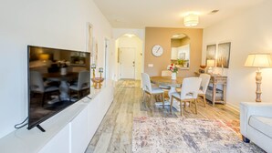 A modern living room with a flat-screen TV, a dining area with a wooden table and chairs, a clock on the wall, and a lamp. - Swim, Golf & Unwind: Barefoot Resort Condo (North Myrtle Beach)