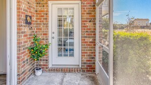 A brick building with a white door, a potted plant, and a view of a tennis court through a window. - Swim, Golf & Unwind: Barefoot Resort Condo (North Myrtle Beach)