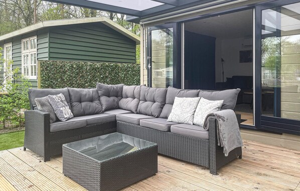 Uma área de estar moderna ao ar livre, com um sofá modular cinza e uma mesa de centro de vidro, sobre um deck de madeira, com uma casa verde ao fundo.