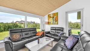 En moderne stue med lædersofaer, et hvidt sofabord og store vinduer med udsigt til en have og omkringliggende huse.