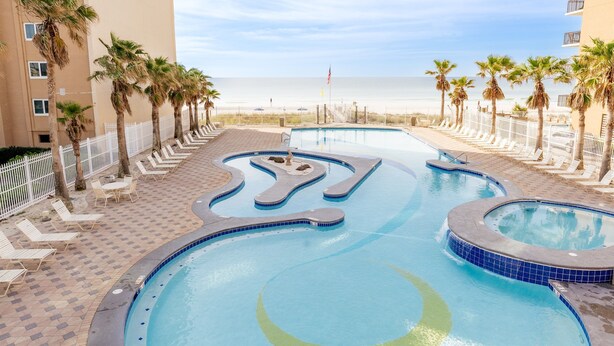 A swimming pool with a unique design, surrounded by palm trees and lounge chairs, with a beach and ocean view in the background. - 3 Pools - Skywalk- 2 Bedroom Updated Condo - Free Daily Activities! (Gulf Shores)