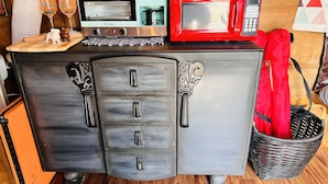 Ein Möbelstück im Vintage-Stil mit Schubladen mit verzierten Griffen, aufgesetzt eine Mikrowelle und ein Toaster, sowie ein Korbwäscher mit rotem Stoff.