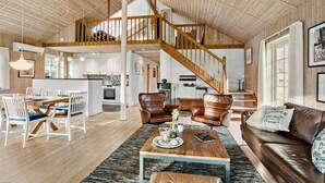 Ein geräumiges Loft mit hoher Decke, Holzbalken, einer zentralen Treppe, einem Essbereich mit weißen Stühlen, einem braunen Ledersofa und einer Küche im Hintergrund.