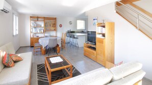 A modern living room with a sofa, coffee table, and a dining area with chairs and a table. There is a kitchen area with a TV and a staircase. - Pretty new and comfortable house between towns and lakes (Voglans)