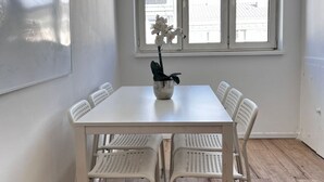 A white dining table with six chairs, a potted plant, and a window with a view of buildings.