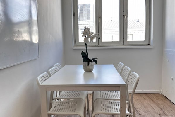 A white dining table with six chairs, a potted plant, and a window with a view of buildings.