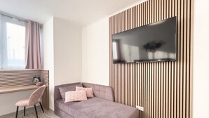 Una habitación moderna con sofá, una silla, televisión montada en una pared de paneles de madera, una ventana con cortinas y un escritorio con lámpara.