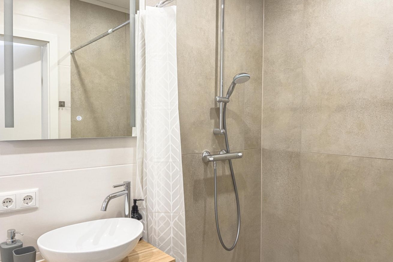 Un baño con lavamanos blanco, un espejo, ducha con cabezal de ducha de mano y un perchero para toallas.