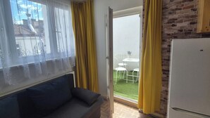 A room with a sofa, a window with curtains, a door leading to an outdoor area with a table and chairs, and a brick wall.