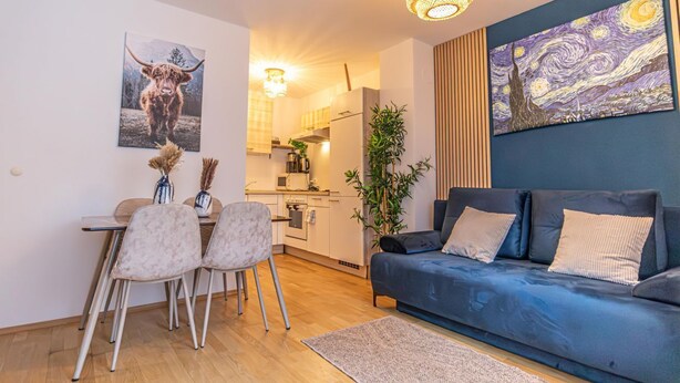 En moderne stue med en blå sofa, et spisebord med stole, et køkken i baggrunden og kunst på væggene.