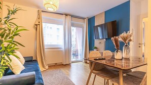 En moderne stue med sofa, spisebord med stole, fjernsyn monteret på væggen og et stort vindue med gardiner.