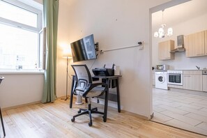 A modern apartment with a wooden floor, a desk with a computer and monitor, a chair, a washing machine, and a kitchen area.