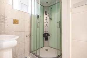 Une cabine de douche fermée par des parois en verre, dotée d’un pommeau de douche à main, d’un panneau de commande fixé au mur et d’un lavabo blanc situé à gauche.