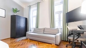 Ein Zimmer mit einer Couch, einem Schreibtisch mit Stuhl, einem Fernseher, einem Schrank und einem Fenster mit Vorhängen.