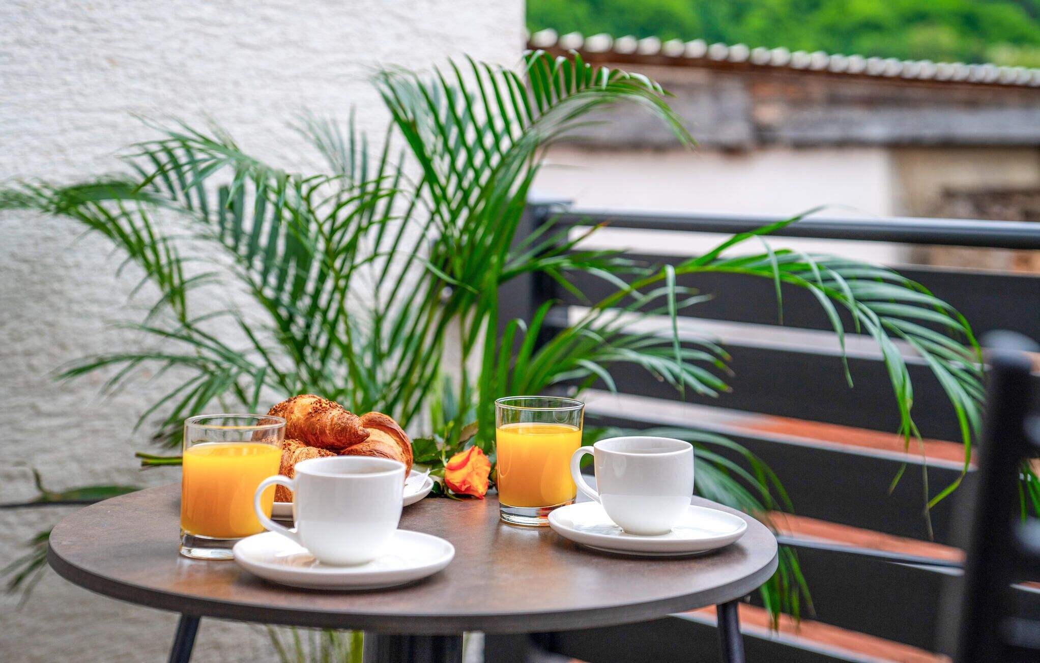 Een ronde tafel met twee koffiekopjes, twee glaasjes sinaasappelsap en een croissant op een balkon, met een witte muur en planten op de achtergrond.