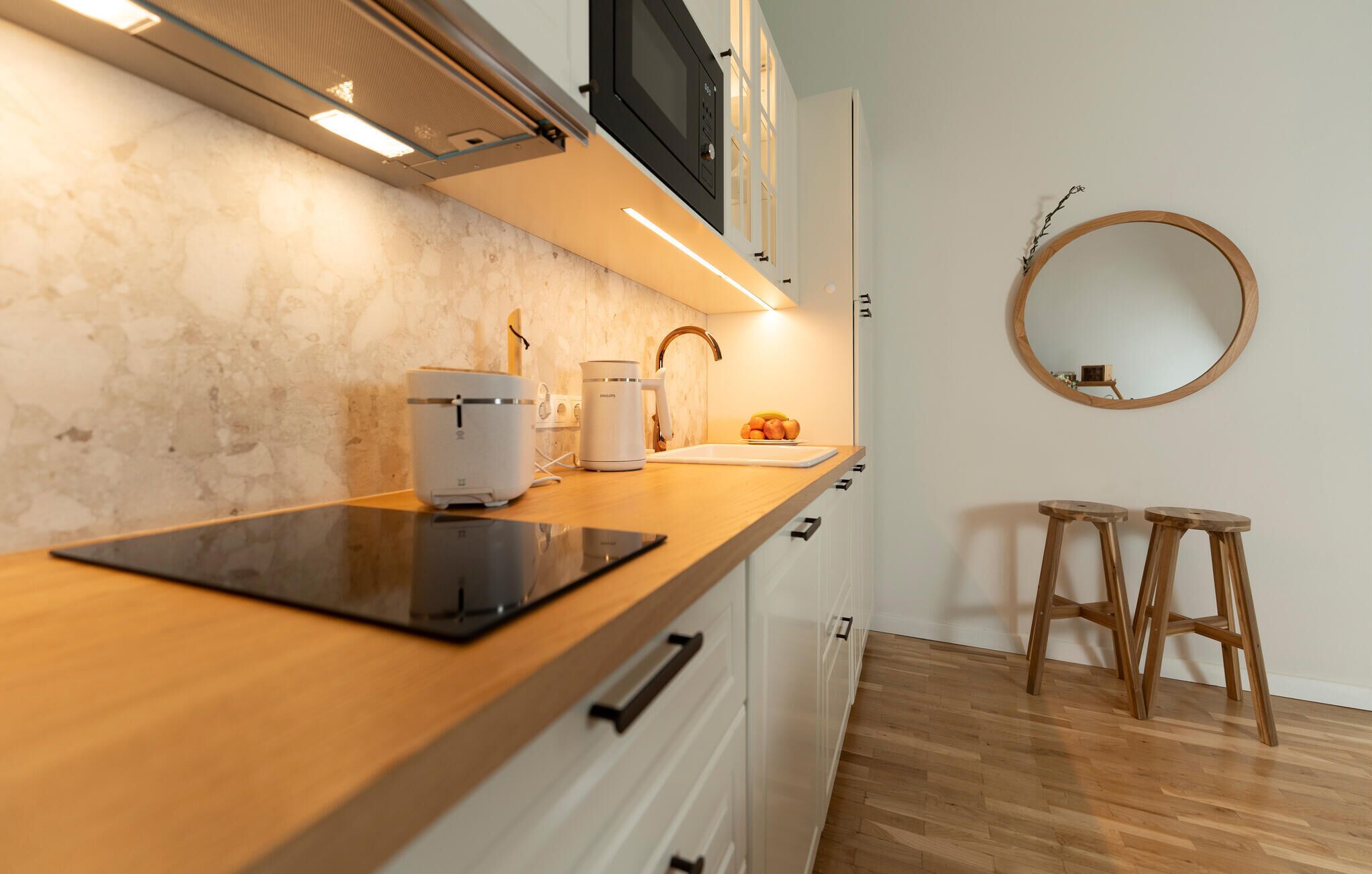 A modern kitchen with a wooden countertop, a built-in microwave, and a sink. There are two wooden stools and a round mirror on the wall.