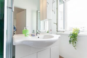 Ein Badezimmer mit einem weißen Waschbecken, einem grünen Seifenspender und einem Fenster mit Blick auf Gebäude.