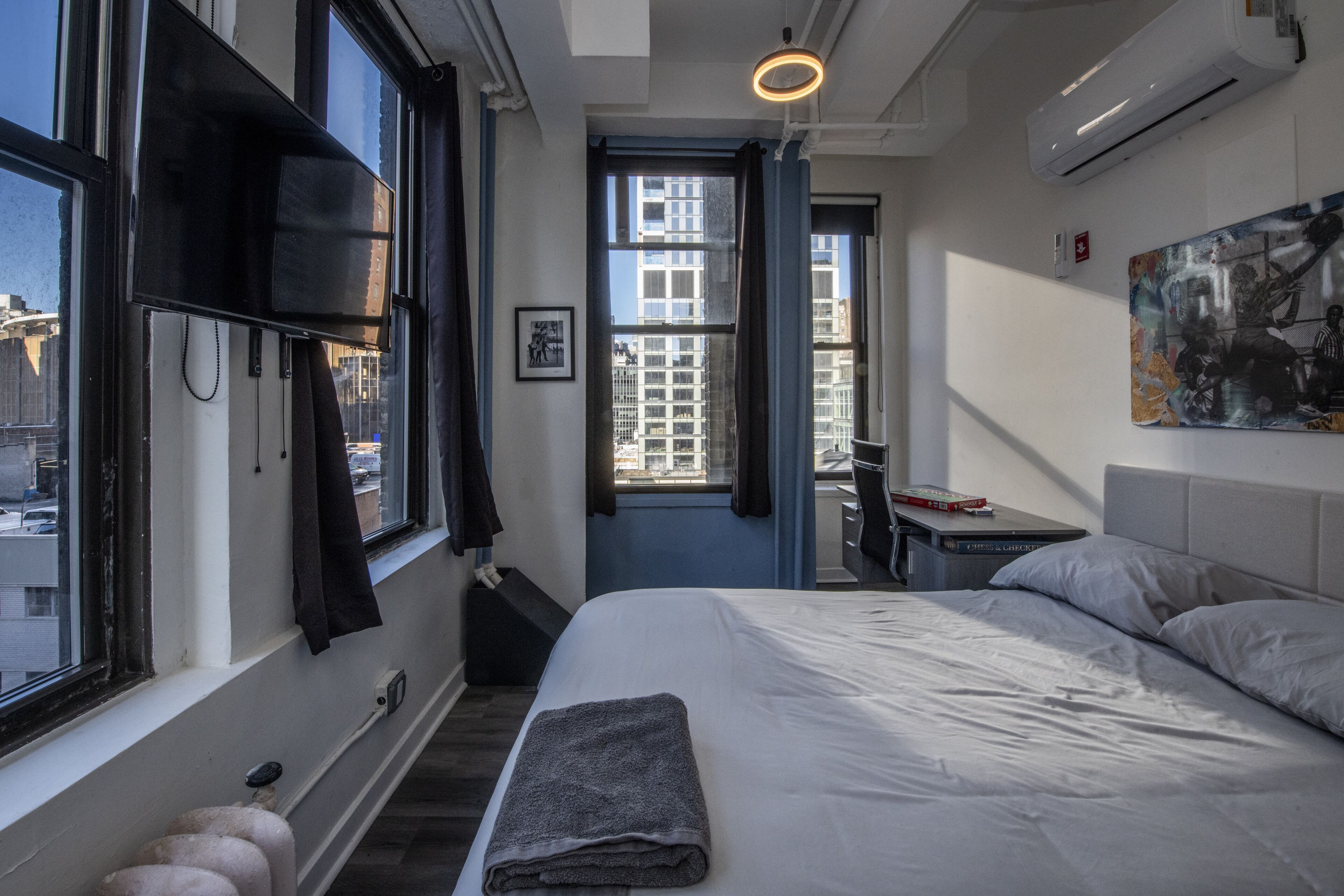 A modern hotel room with a large bed, a flat-screen TV mounted on the wall, a small desk with a chair, and a window offering a city view.