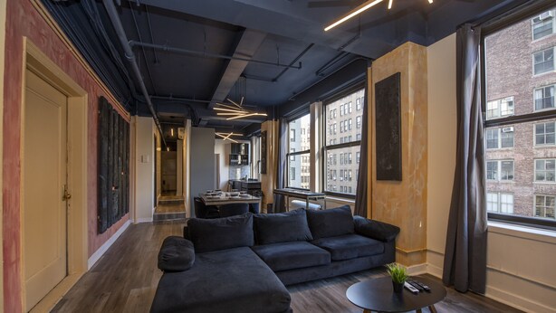 Een moderne woonkamer met een hoekbank, een klein rond tafeltje en uitzicht op de stad door grote ramen.