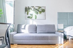 A modern living room with a grey sofa, a wall-mounted picture of eucalyptus leaves, and a bed with a headboard.