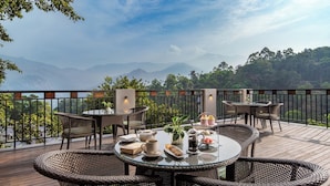 Una terraza con mobiliario de mimbre, una mesa con tetera, tazas y pasteles, con vistas a un paisaje montañoso.