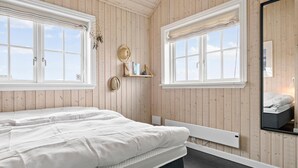 A bedroom with a bed, two windows with curtains, a bedside table, a mirror, and a shelf with books and a hat.