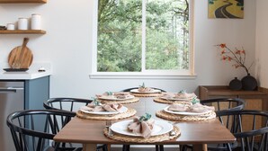 Comedor con mesa de madera dispuesta para seis personas, rodeada de sillas negras. Se ve una ventana con vistas a árboles, y hay estanterías con frascos y un tablón de madera en la pared.