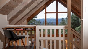 Una habitación con techo inclinado, un escritorio con libros, una silla negra y vistas a montañas a través de amplios ventanales.
