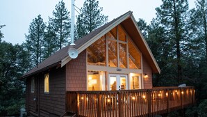 Una cabaña con techo inclinado, ventanas grandes y una terraza con luces colgantes.