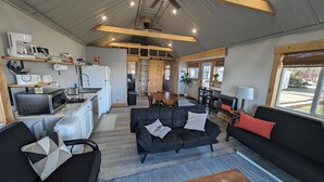 A modern kitchen with a built-in oven, microwave, and coffee maker. There is a dining area with a wooden table and chairs, and a black sofa with cushions. A window provides a view of the outside.