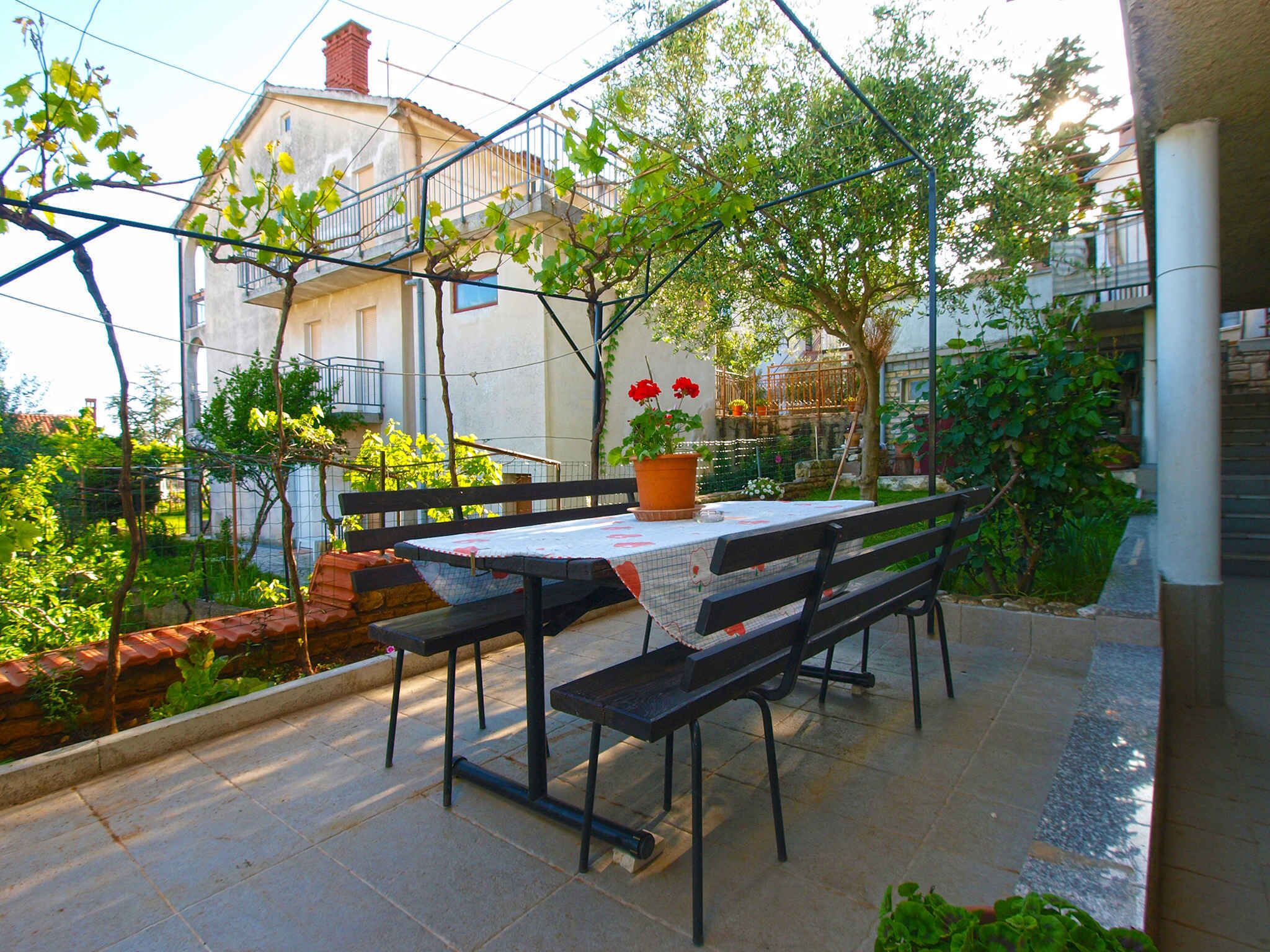 An outdoor patio with a table and benches, surrounded by greenery and a metal fence.