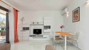 Een moderne woonkamer met een televisie, witte kasten, een eettafel met stoelen en een bank.