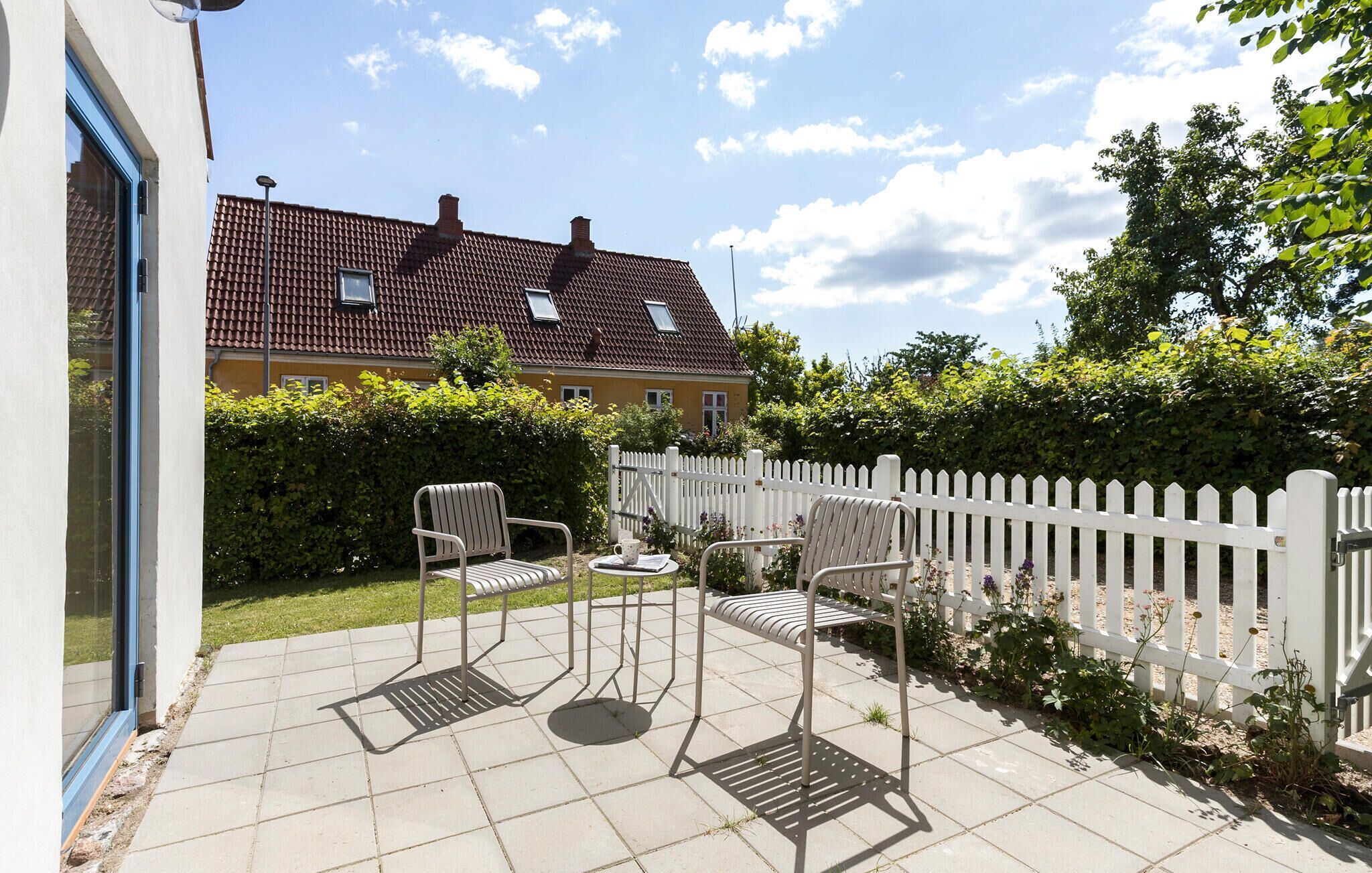Eine Terrasse mit zwei Stühlen und einem Tisch, ein weißer Zierzaun und im Hintergrund ein Haus mit rotem Ziegeldach.