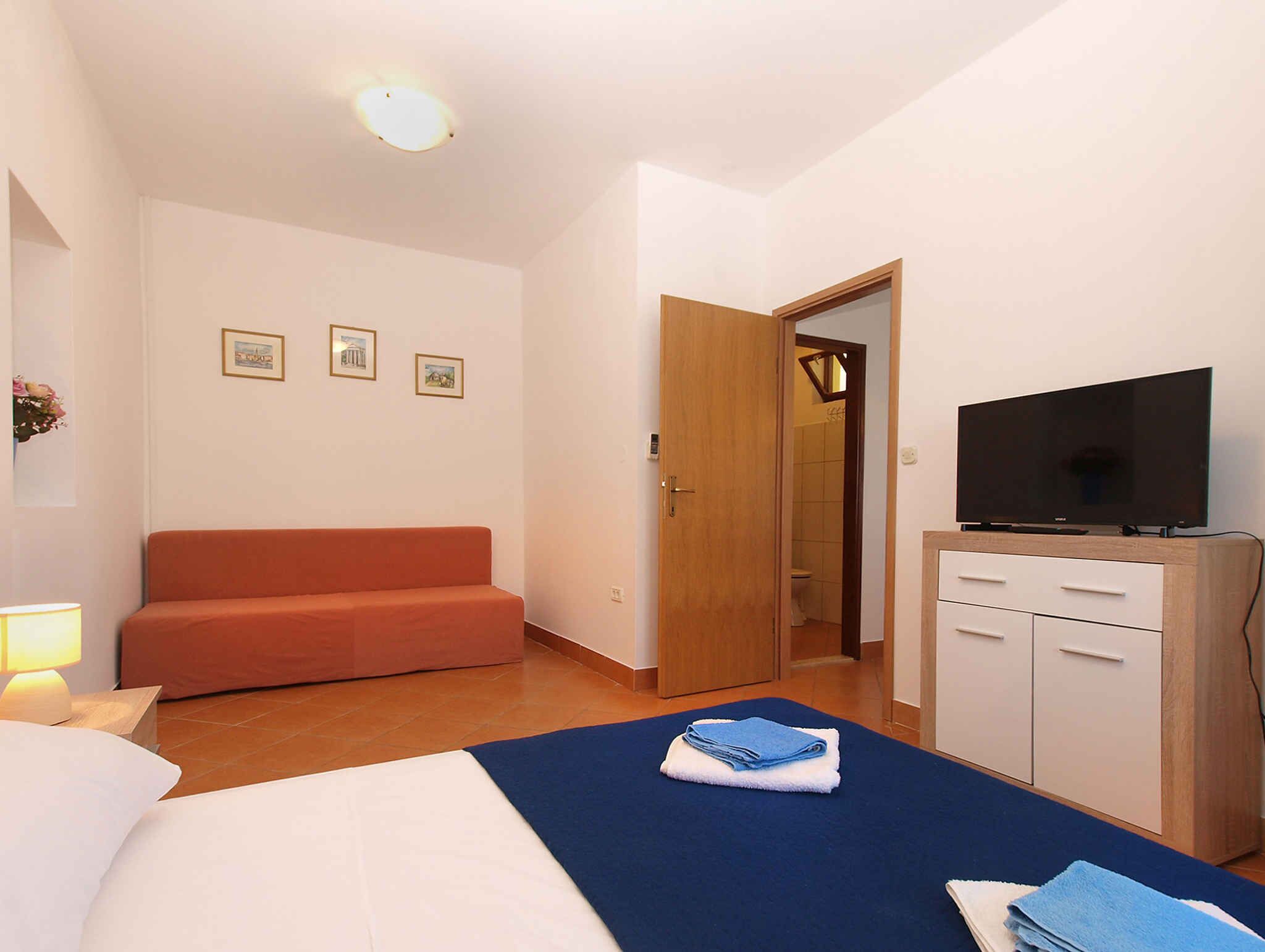 Habitación de hotel con cama, sofá, televisor, armario y baño visible a través de una puerta abierta.