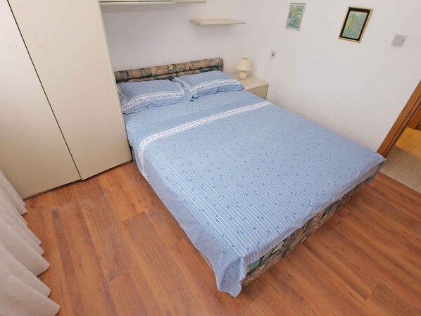 Un lit simple avec une couvre-lit bleue et des oreillers assortis, une table de chevet avec une lampe, et une petite étagère au-dessus du lit.