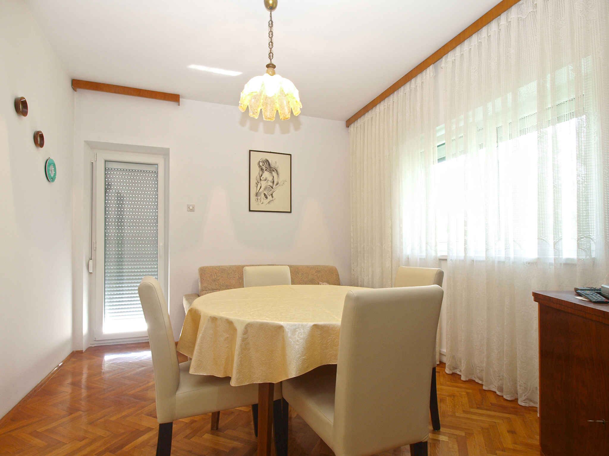 Un comedor con una mesa redonda, sillas, una araña y una puerta con persianas, además de una ventana con cortinas ligeras.