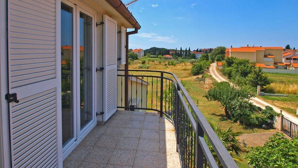 A balcony with a tiled floor and a metal railing, overlooking a rural landscape with houses and greenery.