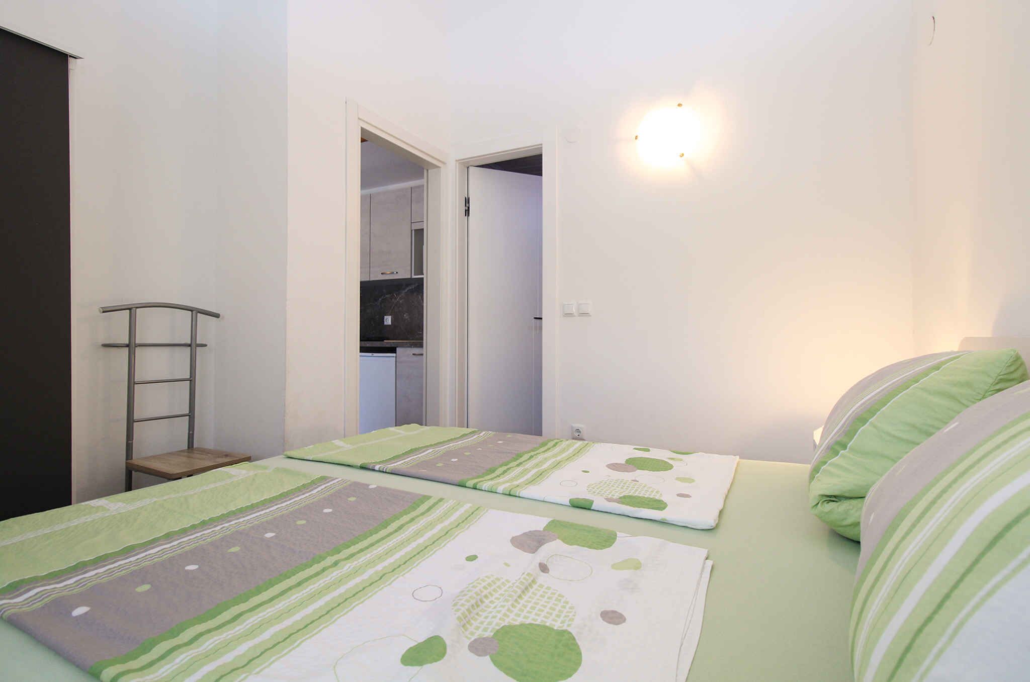 Une petite pièce bien éclairée, avec deux lits, une chaise en bois et une cuisine visible à travers une porte ouverte.