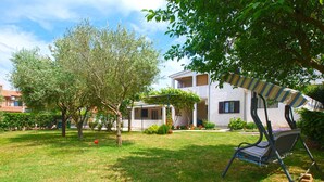 A residential area with a grassy lawn, trees, a patio with a striped umbrella, and a building with balconies. - Holiday flat with air conditioning (Valbandon)