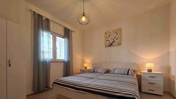 A bedroom with a large bed, two bedside tables, a hanging light fixture, a window with curtains, and a wall-mounted artwork. - Holiday flat with air conditioning (Valbandon)