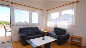 A living room with a black leather sofa, a white coffee table, and a wooden floor. There are two windows with white curtains and a view of a building outside.