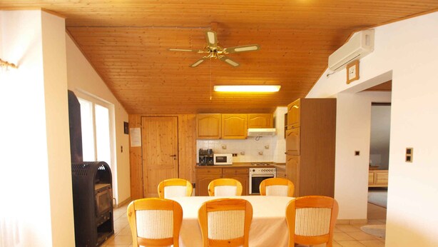 A dining area with a wooden ceiling, a ceiling fan, and a kitchen in the background.