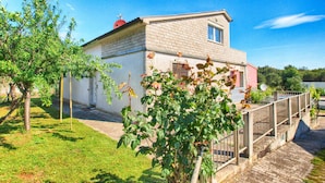 A two-story house with a brick facade, a small balcony, and a garden area with trees and a flower bush.