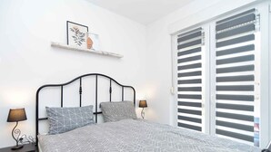 A bedroom with a bed, two bedside lamps, a shelf with a framed picture, and a window with blinds.