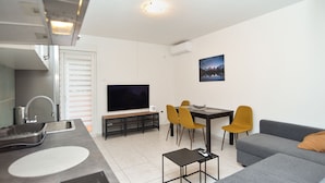 A modern living room with a flat-screen TV, a dining area with yellow chairs, a grey sofa, and a kitchen area with a sink and stove.