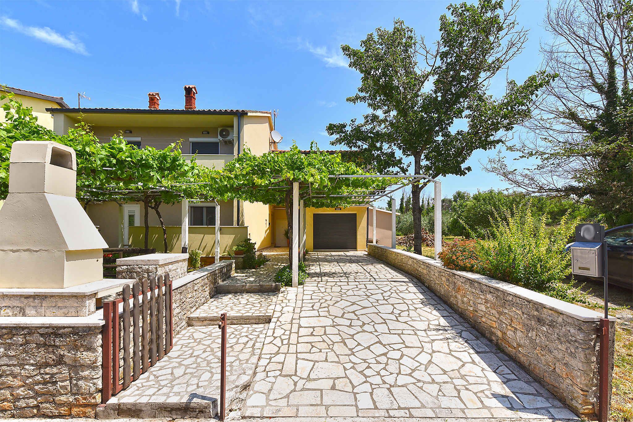 Un chemin pavé de pierres mène à un bâtiment jaune doté d’un garage, le tout étant entouré de verdure et surplombé par un ciel dégagé.