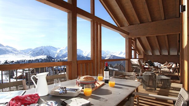 A dining area with a table set for breakfast, a view of snow-capped mountains, and wooden beams in the ceiling.