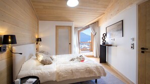 A cozy bedroom with a wooden ceiling, a bed with a fluffy blanket, a bedside lamp, a small table with a book, and a view of a mountain through a window.